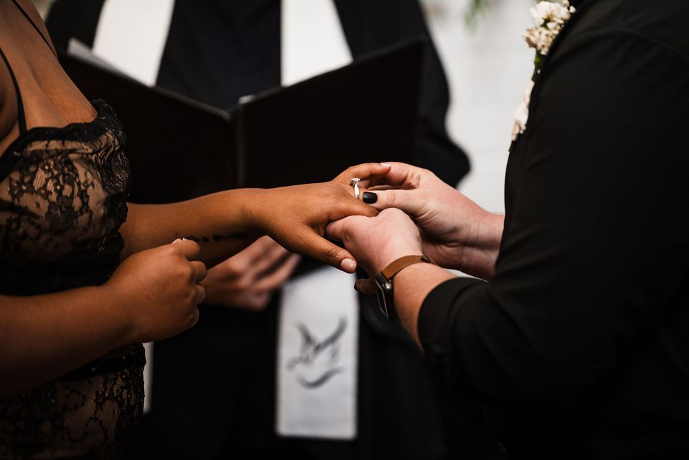 Two hands exchanging rings during an intimate wedding ceremony with an officiant in the background.