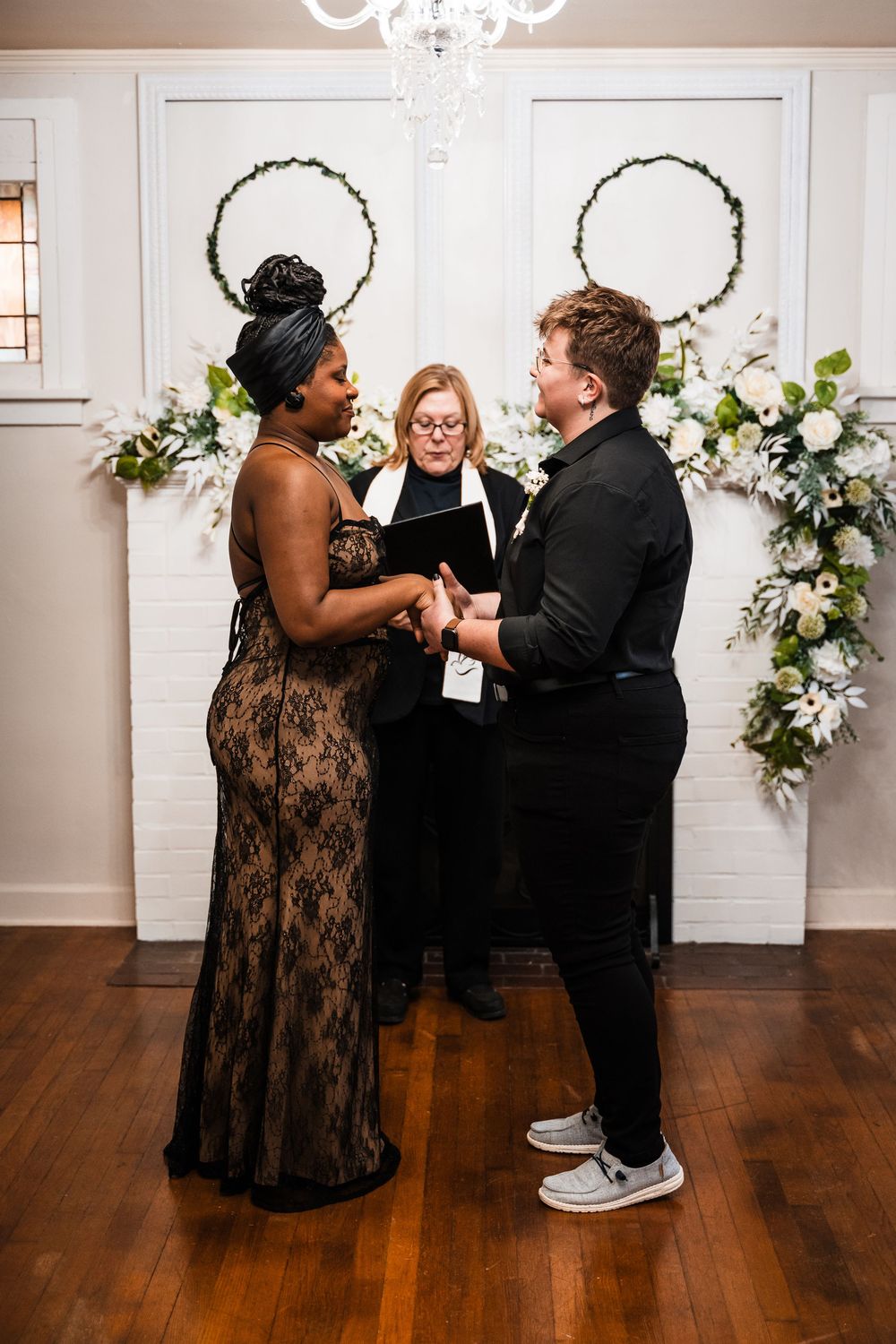Indoor wedding ceremony with couple holding hands in front of officiant and floral arrangements.