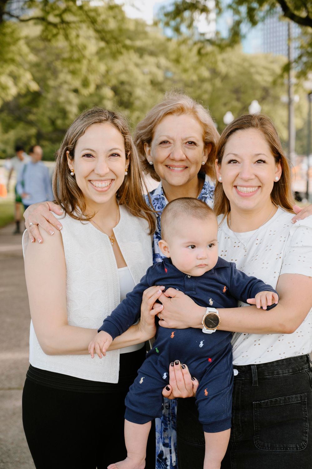 Three people in white tops gather together outdoors with a baby wearing a navy outfit on a sunny day.