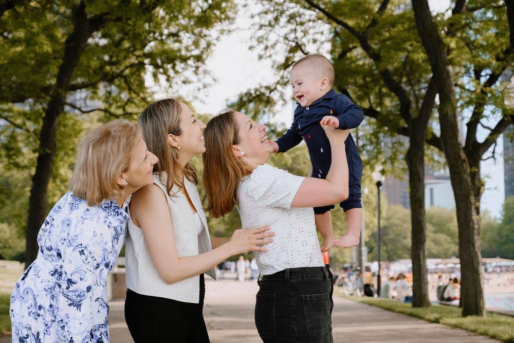 Three people spend time together outdoors lifting up a baby while standing near trees on a sunny day.