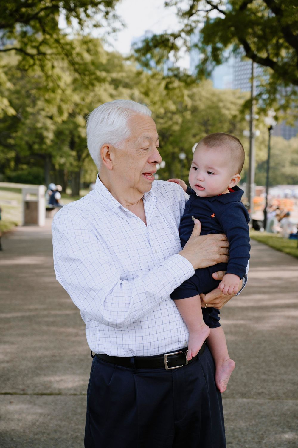 An elderly man in a white button-down shirt holds a small baby wearing navy blue clothing outdoors on a sunny day.
