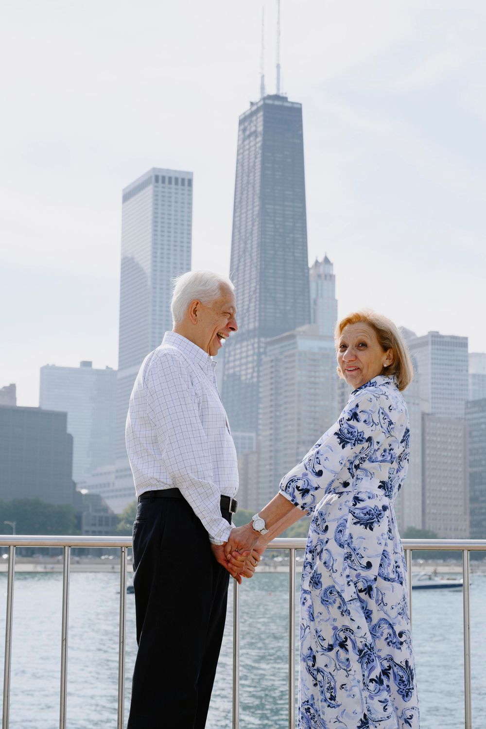 A couple holding hands on a waterfront with the Chicago skyline and John Hancock building in the background.