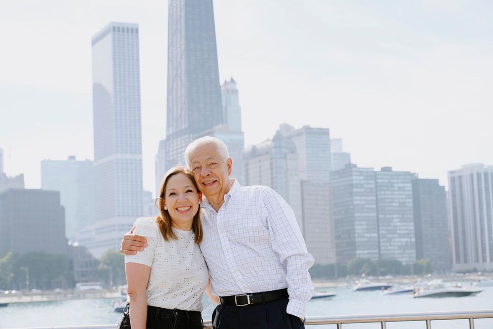 Two people stand together smiling by the Chicago waterfront with tall city skyscrapers in the background.