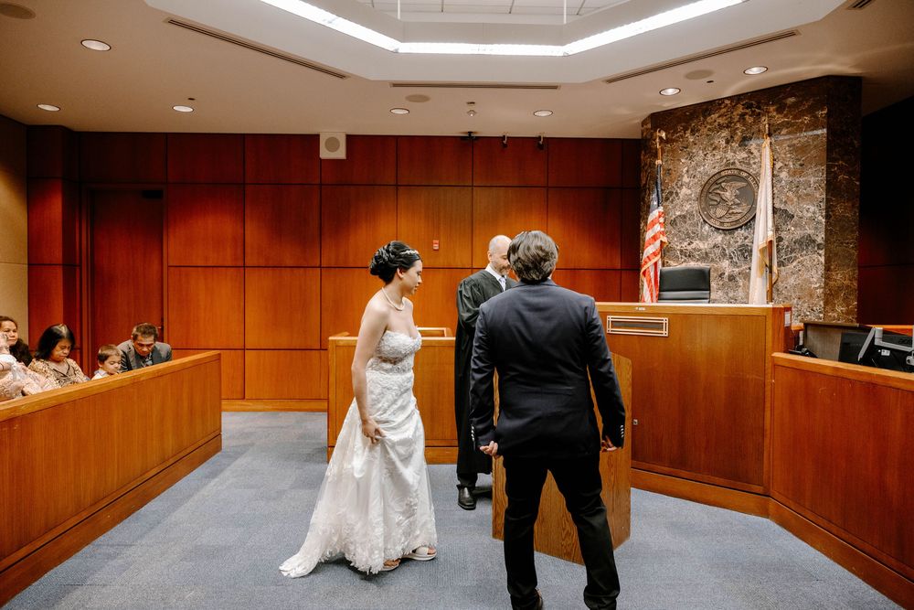 A couple stands before a judge in a wood-paneled courtroom with stone accents during their civil ceremony.