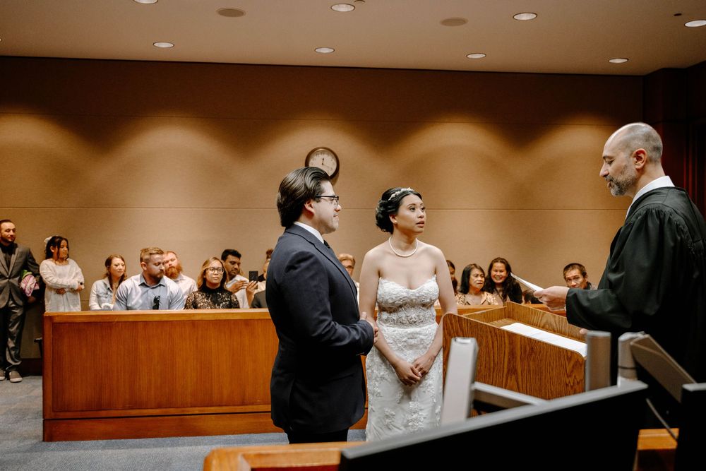 Two people in wedding attire stand before an official during an indoor courthouse marriage ceremony.