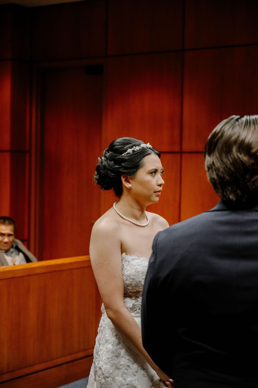 A bride in a lace wedding dress and pearl necklace exchanges vows in a wood-paneled courtroom.