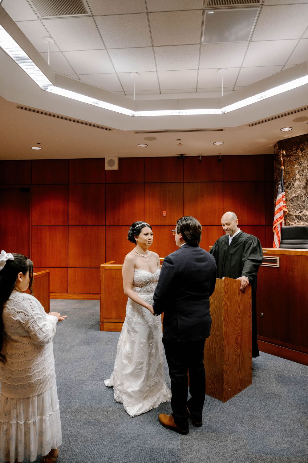 A wedding ceremony taking place in a wood-paneled courtroom with decorative ceiling lights.