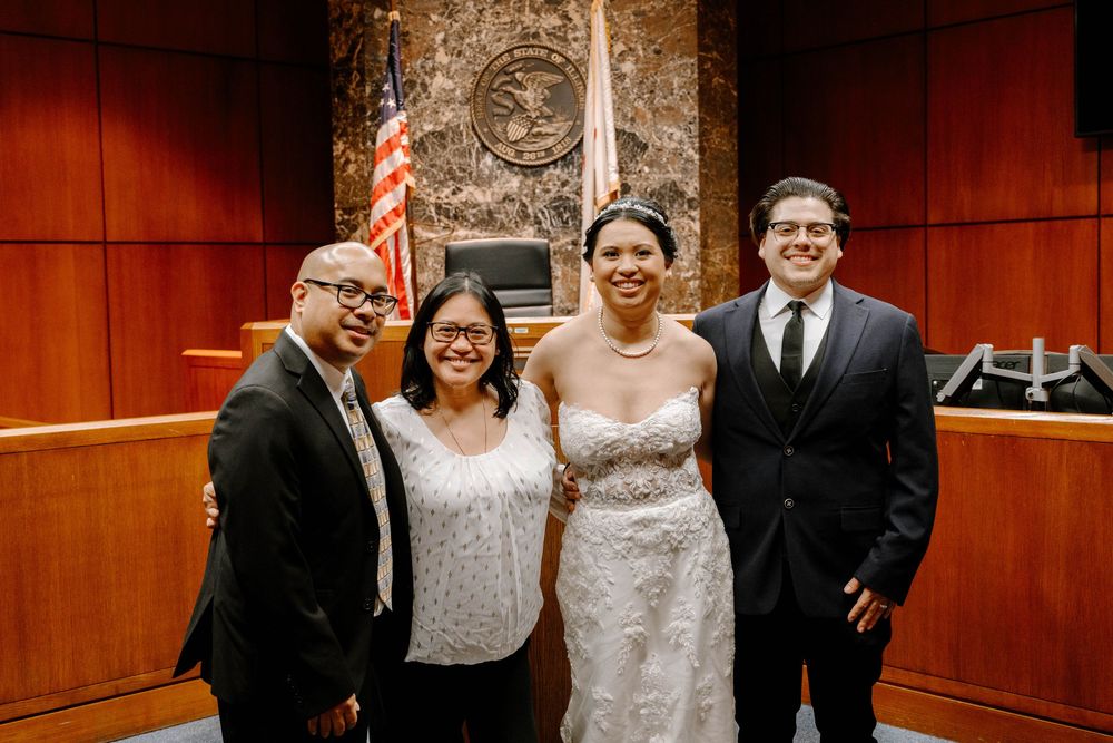 A group of four people stand together smiling in front of a wood-paneled courtroom wall with an American flag.