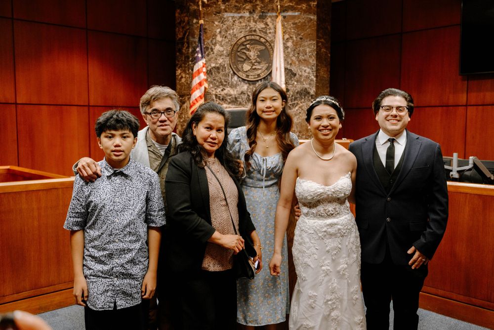 A group of six people pose together in formal attire inside a wood-paneled courtroom with a large seal on the wall.
