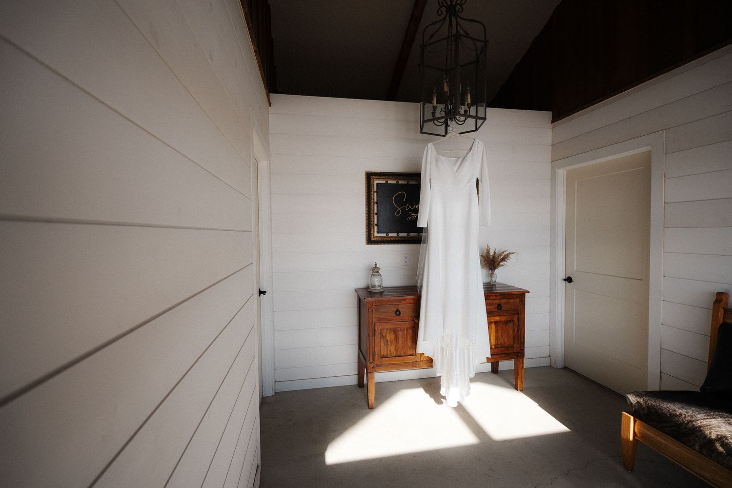 A white wedding dress hangs elegantly in a sunlit room with white walls, wooden dresser and vintage chandelier.