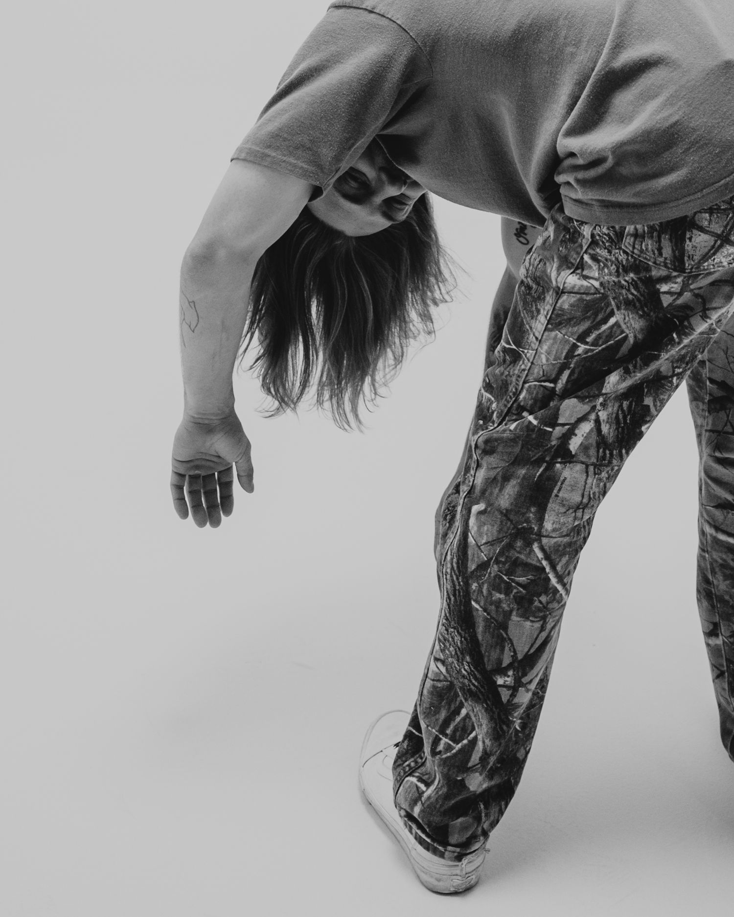 Black and white photo of figure in camouflage pants bending backwards with dark hair falling down against light background.