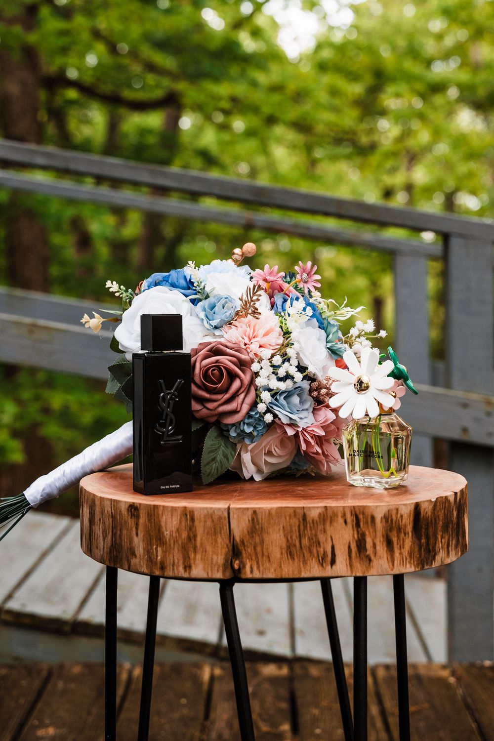 Wedding bouquet with pink and blue flowers displayed on rustic wood slice table with perfume bottle.