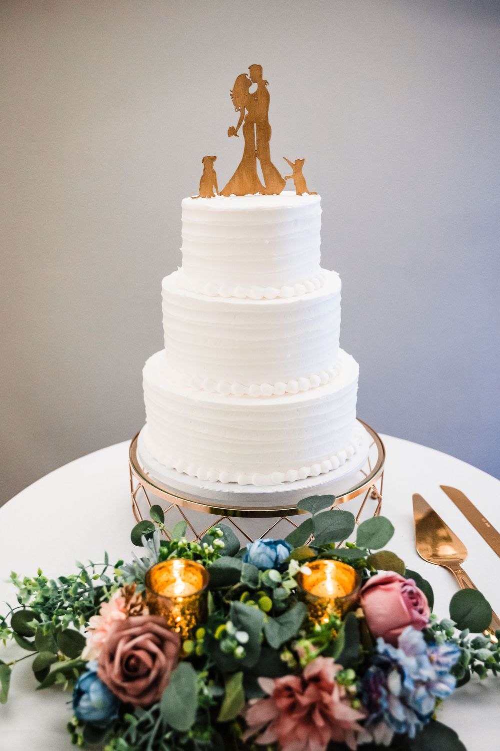 Three-tiered white wedding cake with golden silhouette topper surrounded by floral decor and candles.