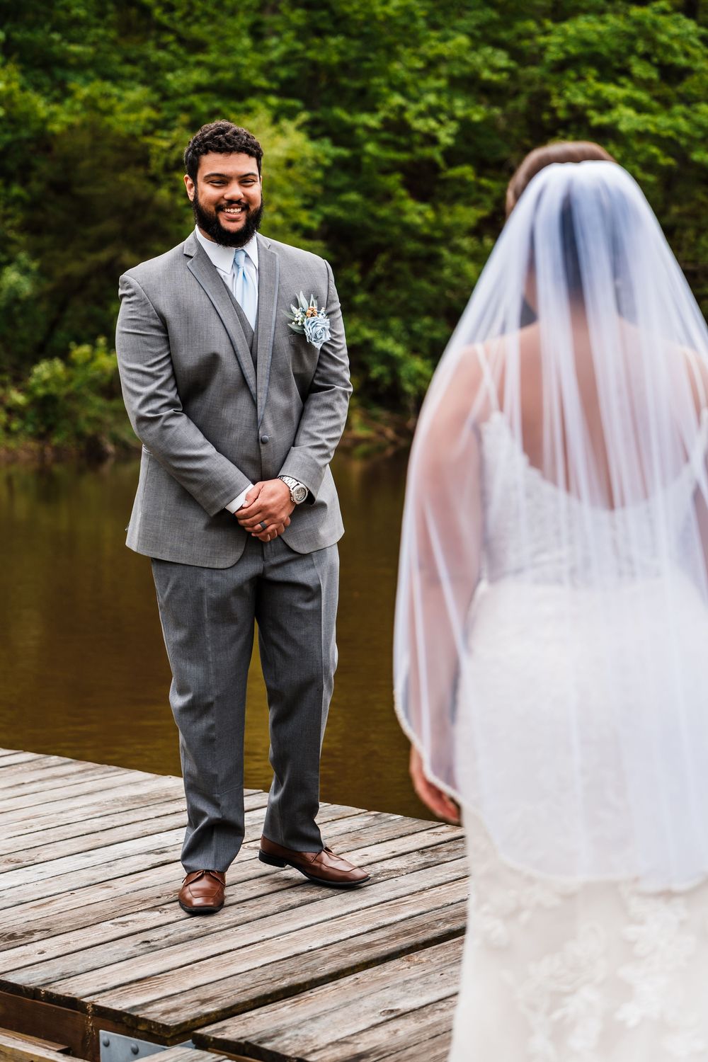 Groom in a grey suit stands on a wooden dock by a lake while viewing his bride in her white wedding veil.