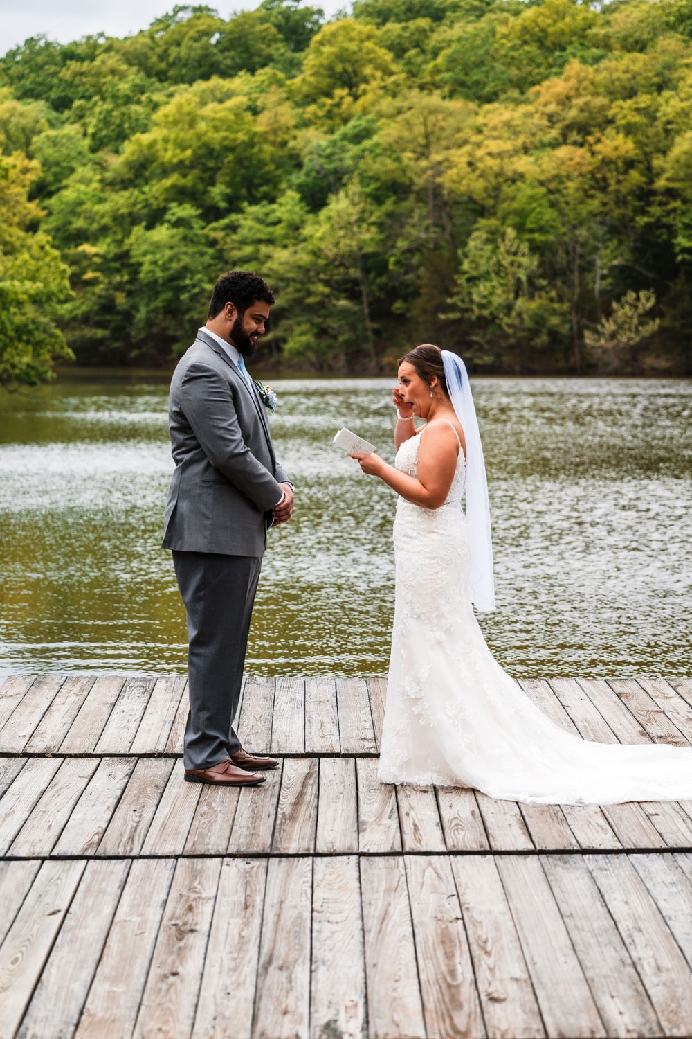 A couple exchanges rings during their wedding ceremony on a wooden dock by a serene lake surrounded by lush trees.
