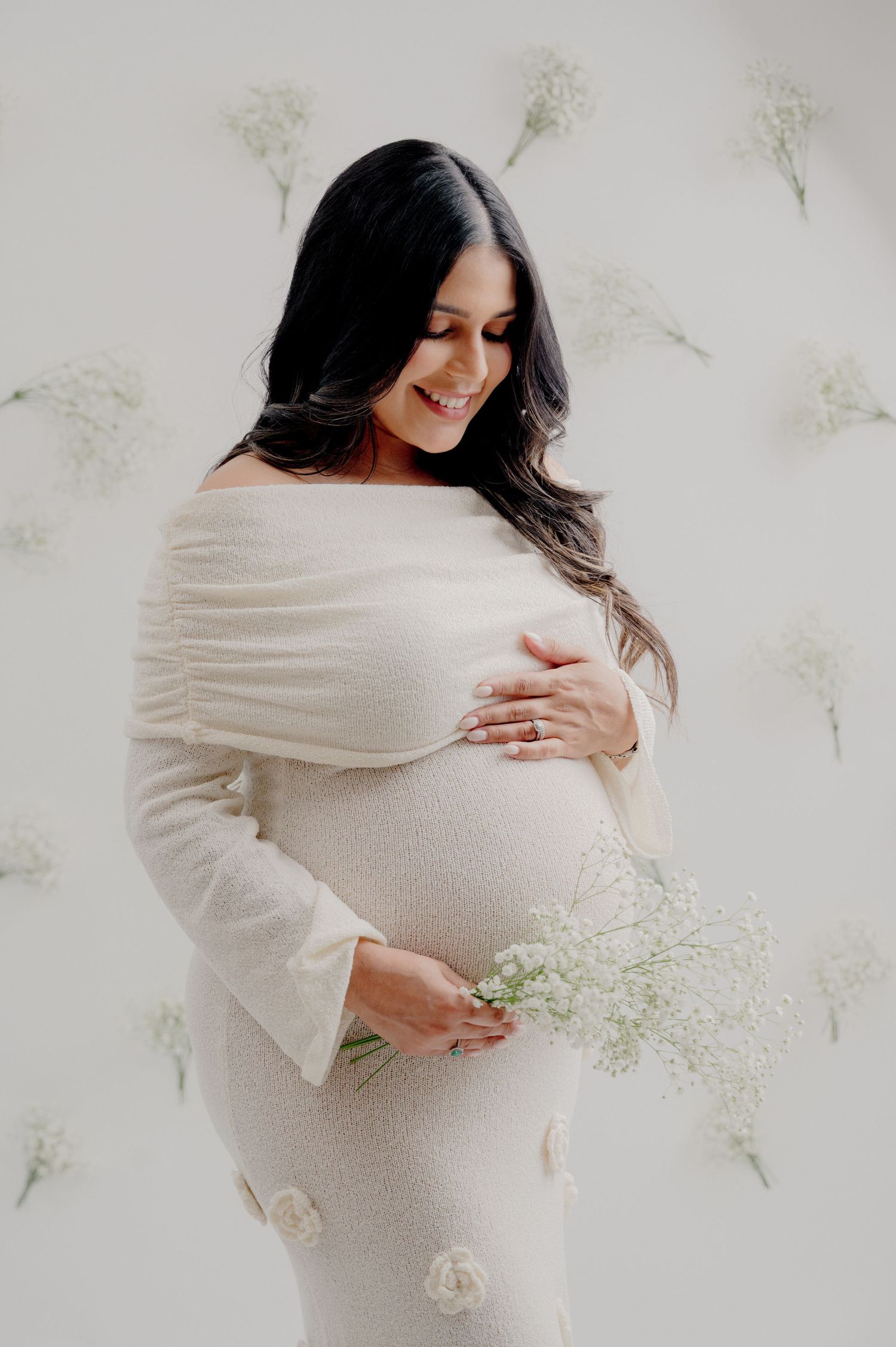 Expectant mother in white flowing dress poses against floral backdrop holding delicate baby's breath flowers.