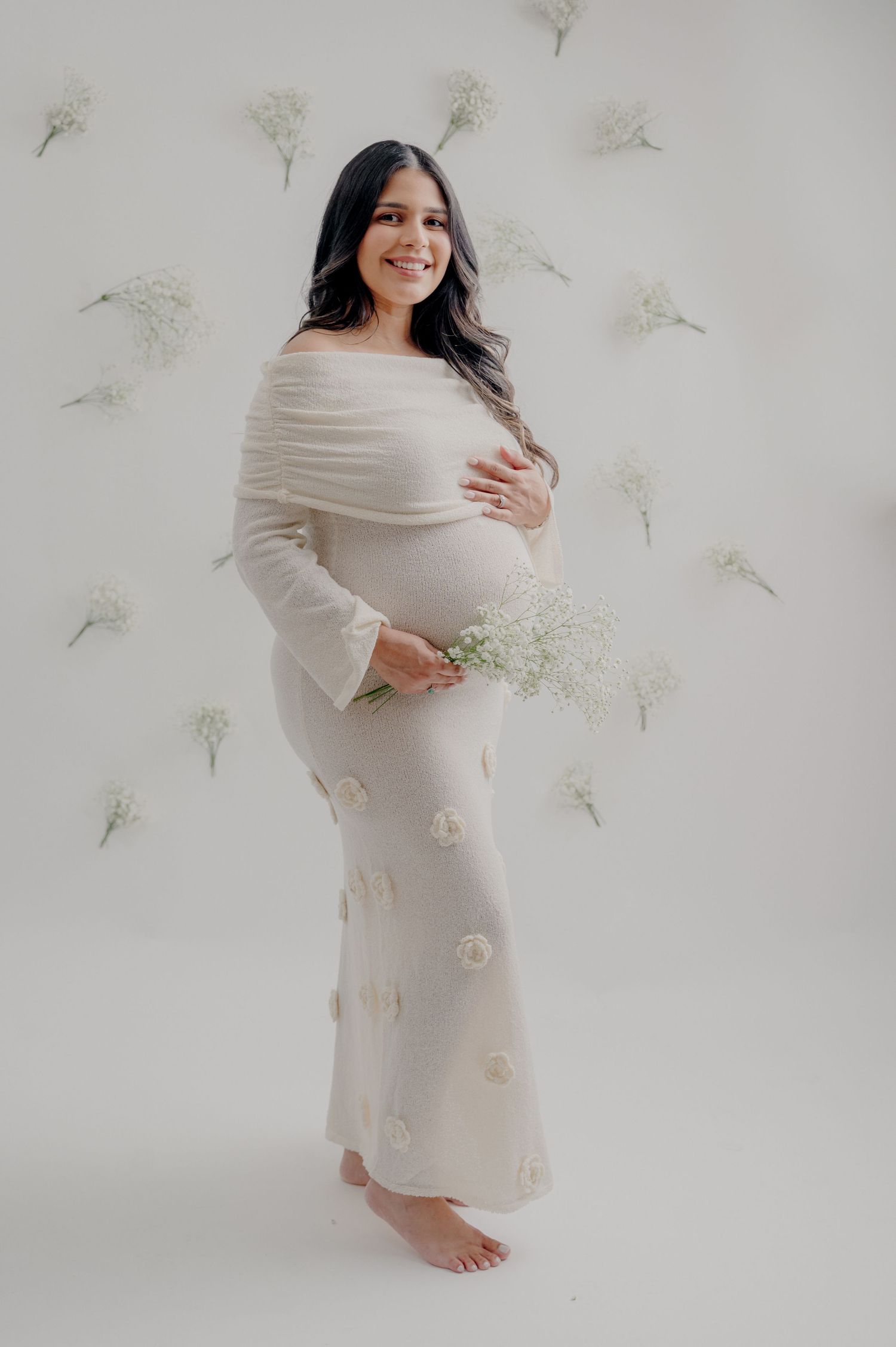Expectant mother wearing a flowing white dress poses barefoot against wall decorated with delicate dandelion designs.
