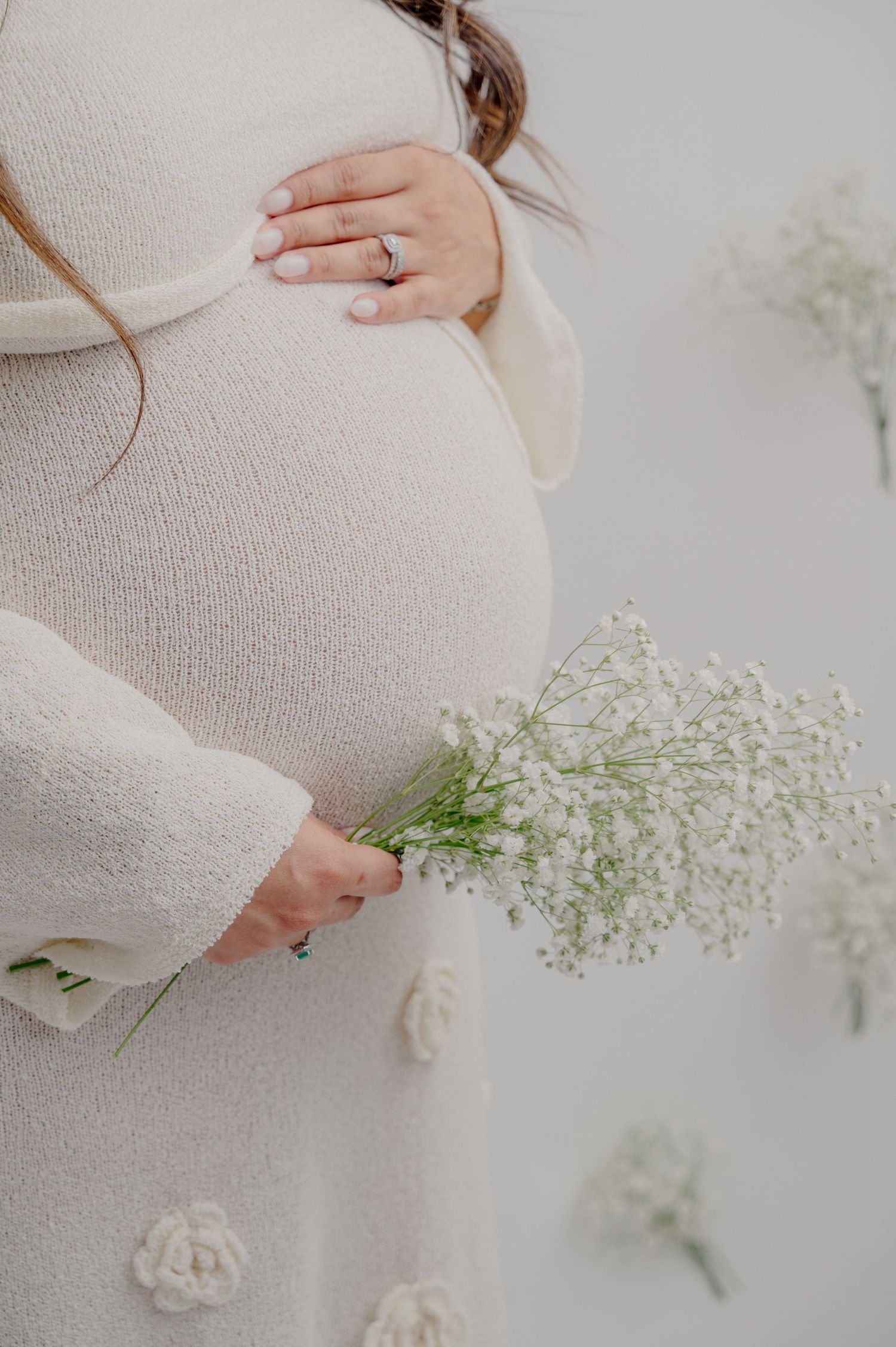 A pregnant woman in a cream sweater holds delicate baby's breath flowers against her belly, with a sparkling ring visible.
