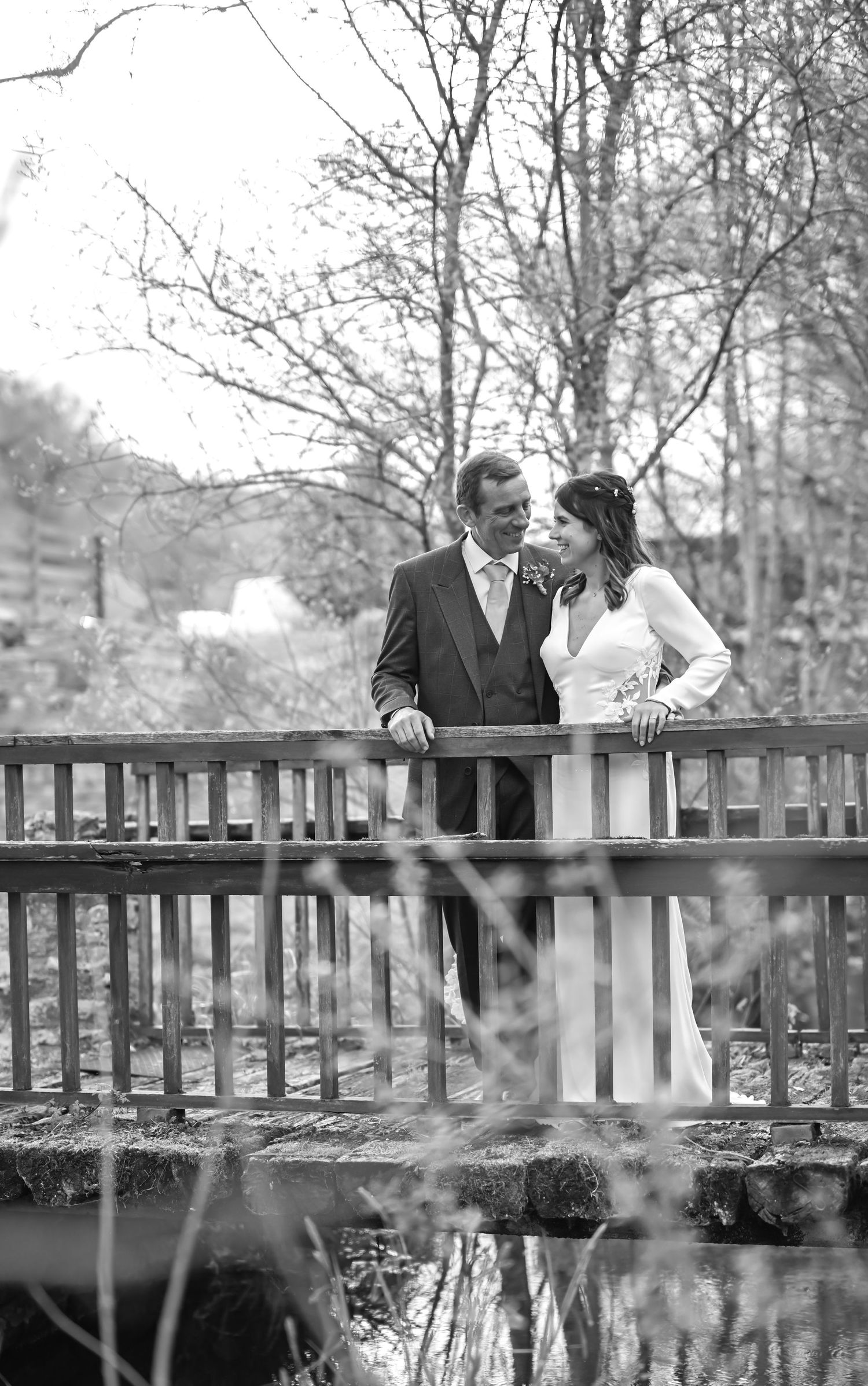 Romantic black and white wedding photo of couple sharing intimate moment on rustic wooden bridge surrounded by lush foliage.