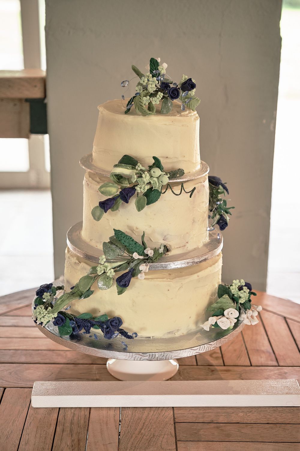 Three-tiered white buttercream wedding cake decorated with fresh blueberries, greenery and small white flowers on a cake stand.
