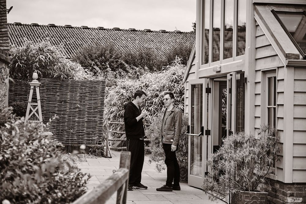 A vintage black and white photograph showing figures standing near a house entrance with a thatched fence in the background.