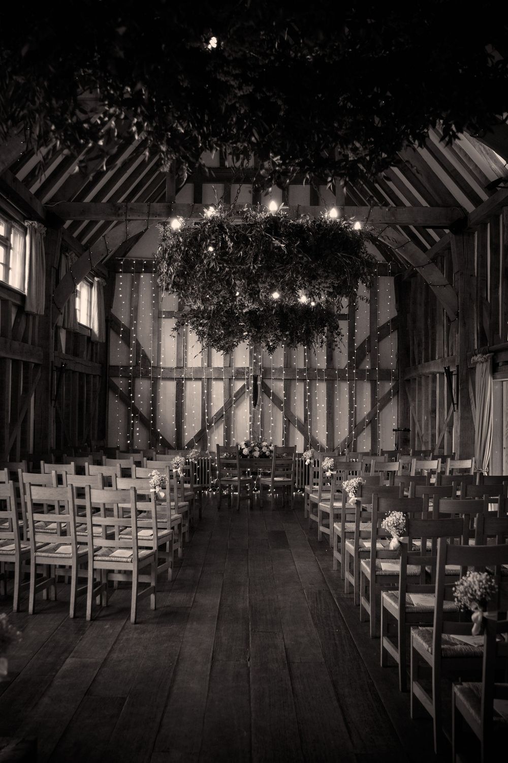 Rustic barn wedding venue interior with rows of chairs and hanging lights beneath exposed wooden beams in black and white.