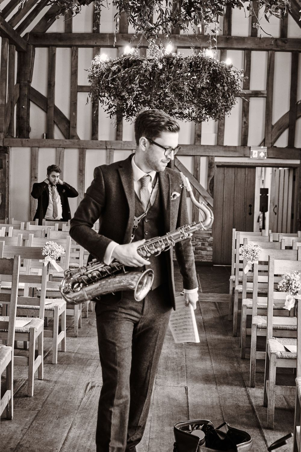 Black and white photo of a saxophone player performing in a rustic wooden barn venue with rows of empty chairs.