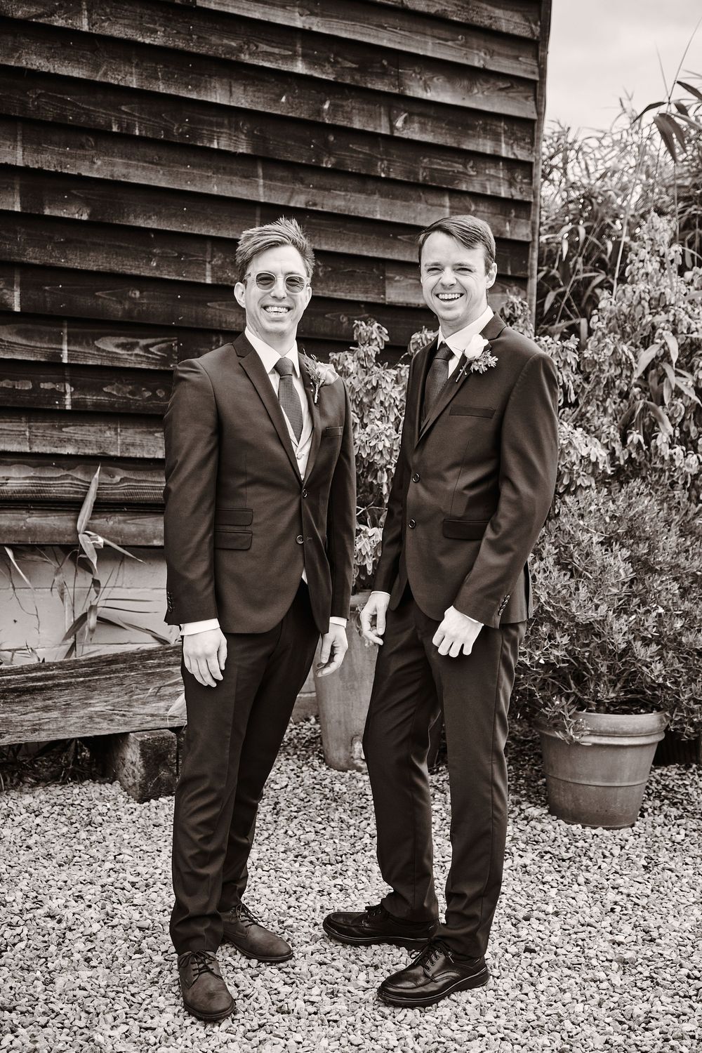 Two men in formal dark suits and boutonnières stand together in front of a rustic wooden wall at an outdoor event.