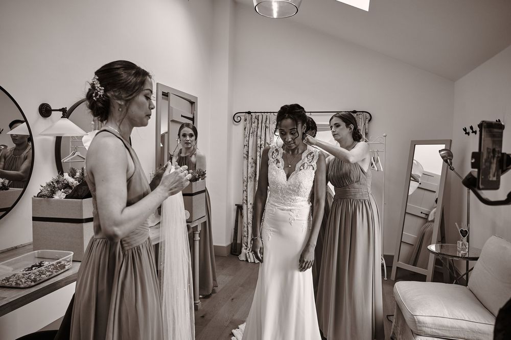 Black and white photo showing bridal preparations with bridesmaids in long dresses helping in a getting ready room.