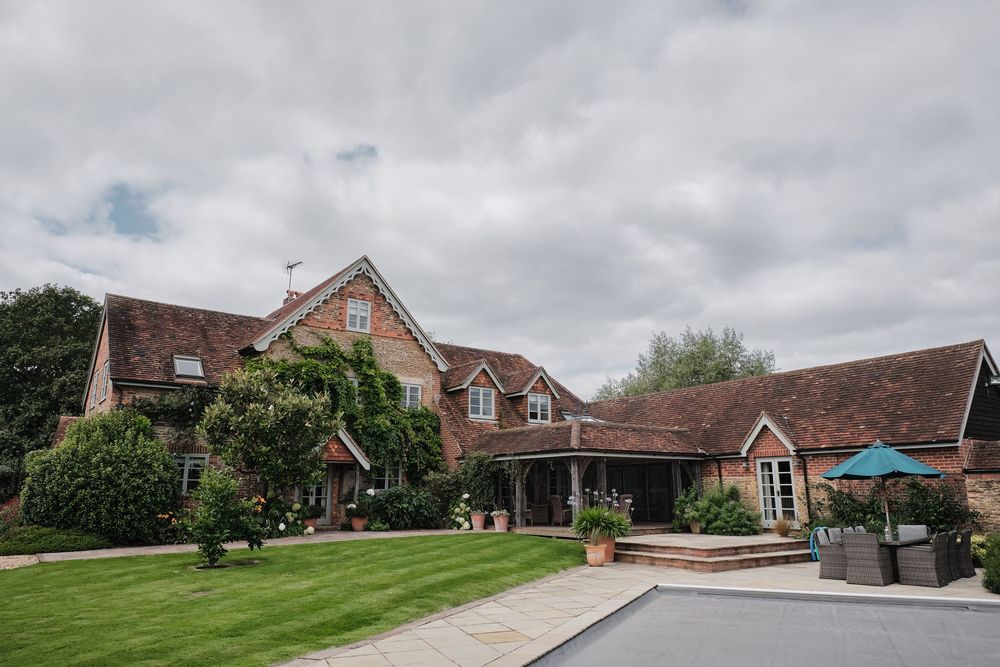 Rustic brick country house with manicured lawn and overhanging greenery against cloudy sky.