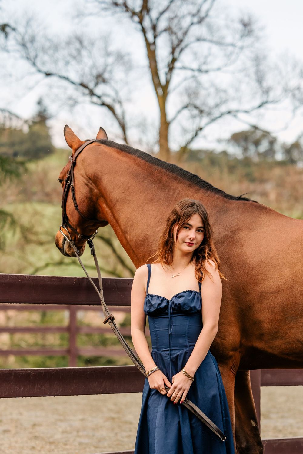 A brown horse stands next to someone wearing a blue denim dress holding a riding crop in an outdoor paddock.
