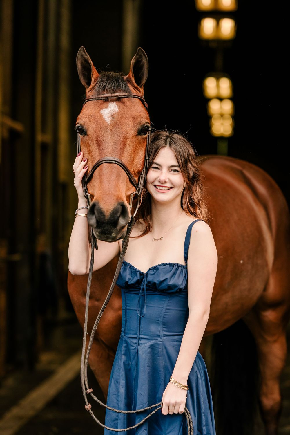 A beautiful chestnut horse with a black bridle stands next to someone in a navy blue dress inside a rustic stable.