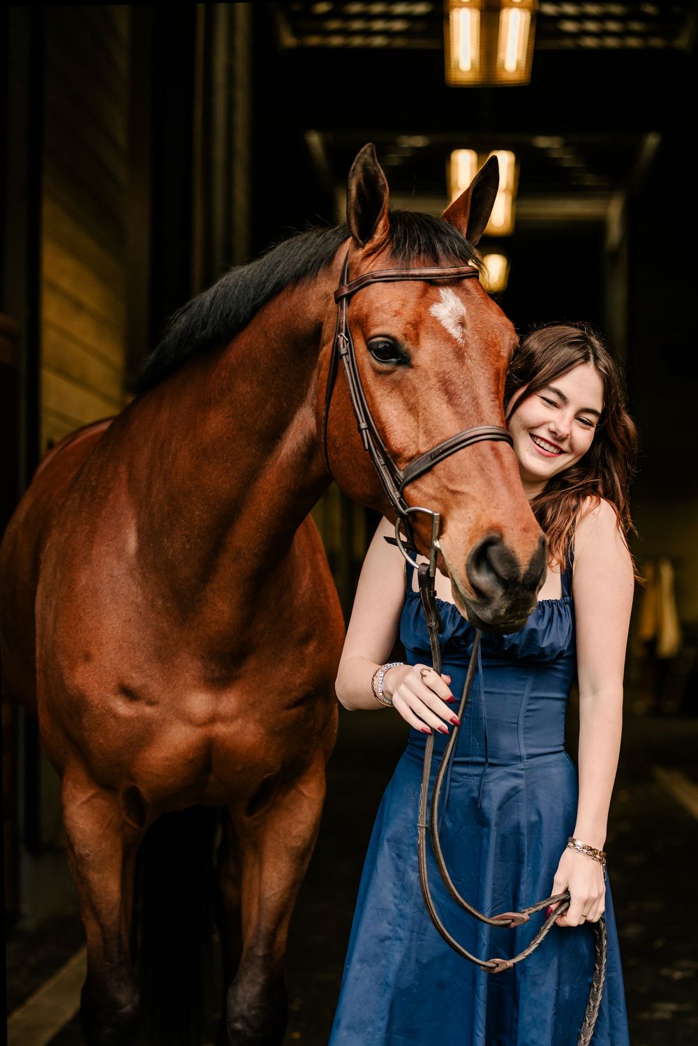 A beautiful bay horse with a dark bridle stands in a barn stall next to someone in a blue denim dress.
