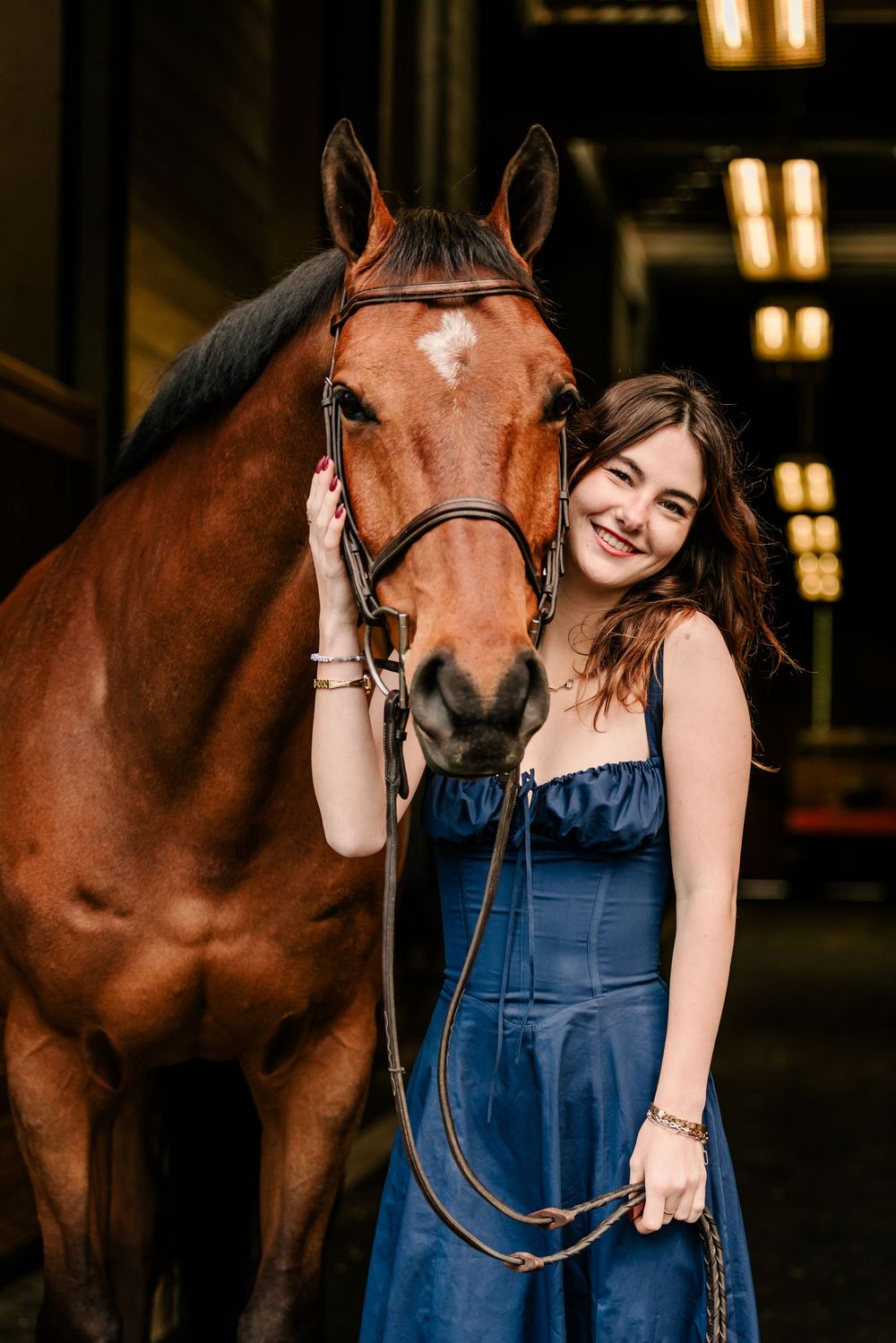 A beautiful bay horse stands next to someone wearing a blue corset dress in what appears to be a barn interior.