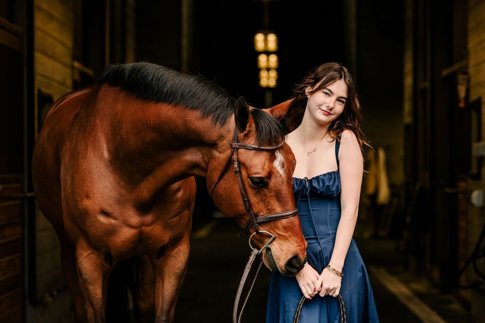 A person in a dark blue dress stands with a chestnut bay horse in a dimly lit stable setting.