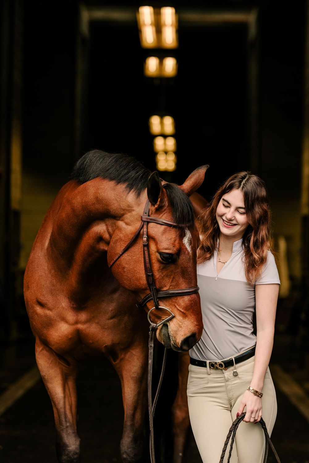 Beautiful chestnut horse and rider share a tender moment in a dimly lit stable with warm overhead lighting.
