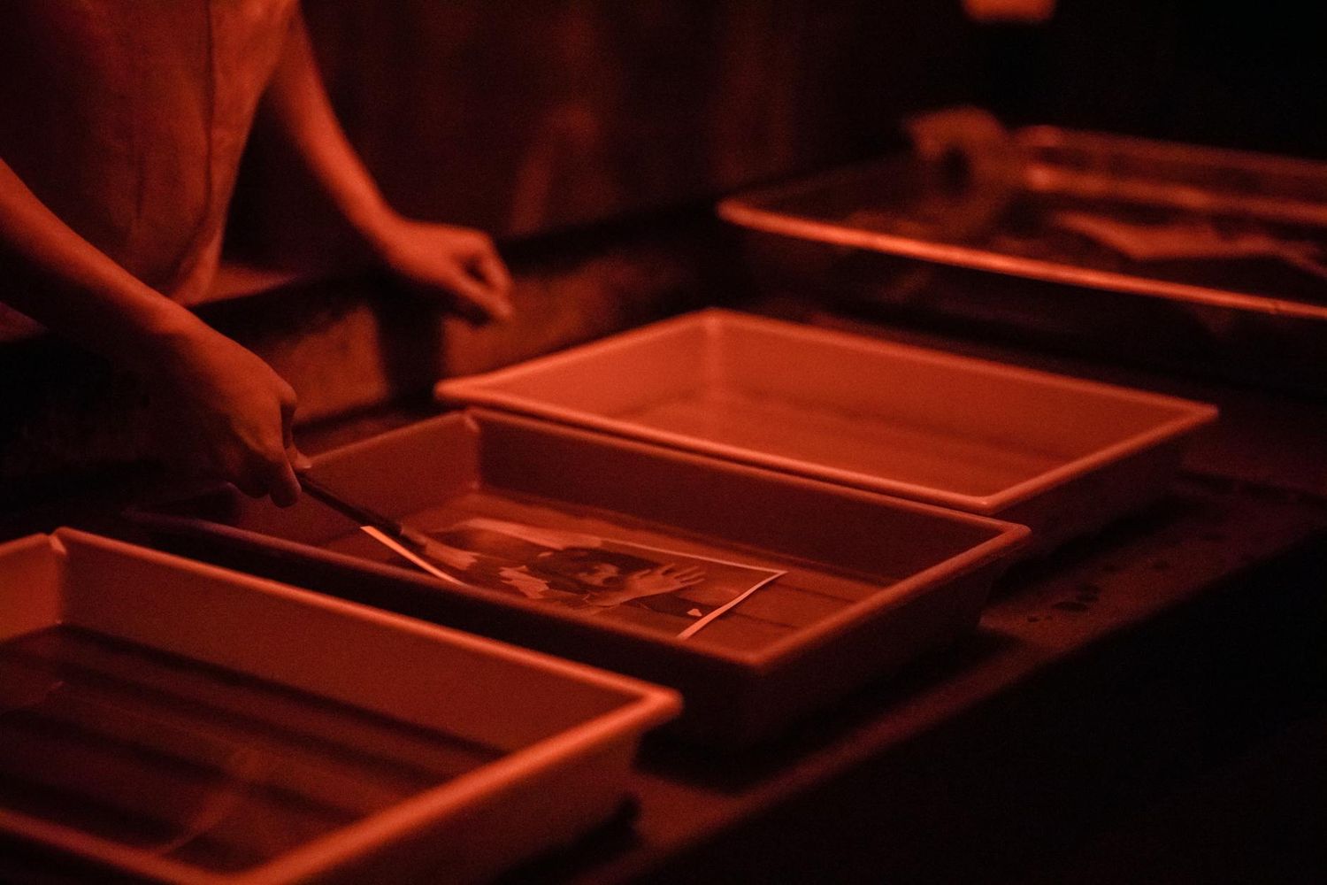 Professional darkroom photography development process showing orange-lit trays with photo paper and chemicals.