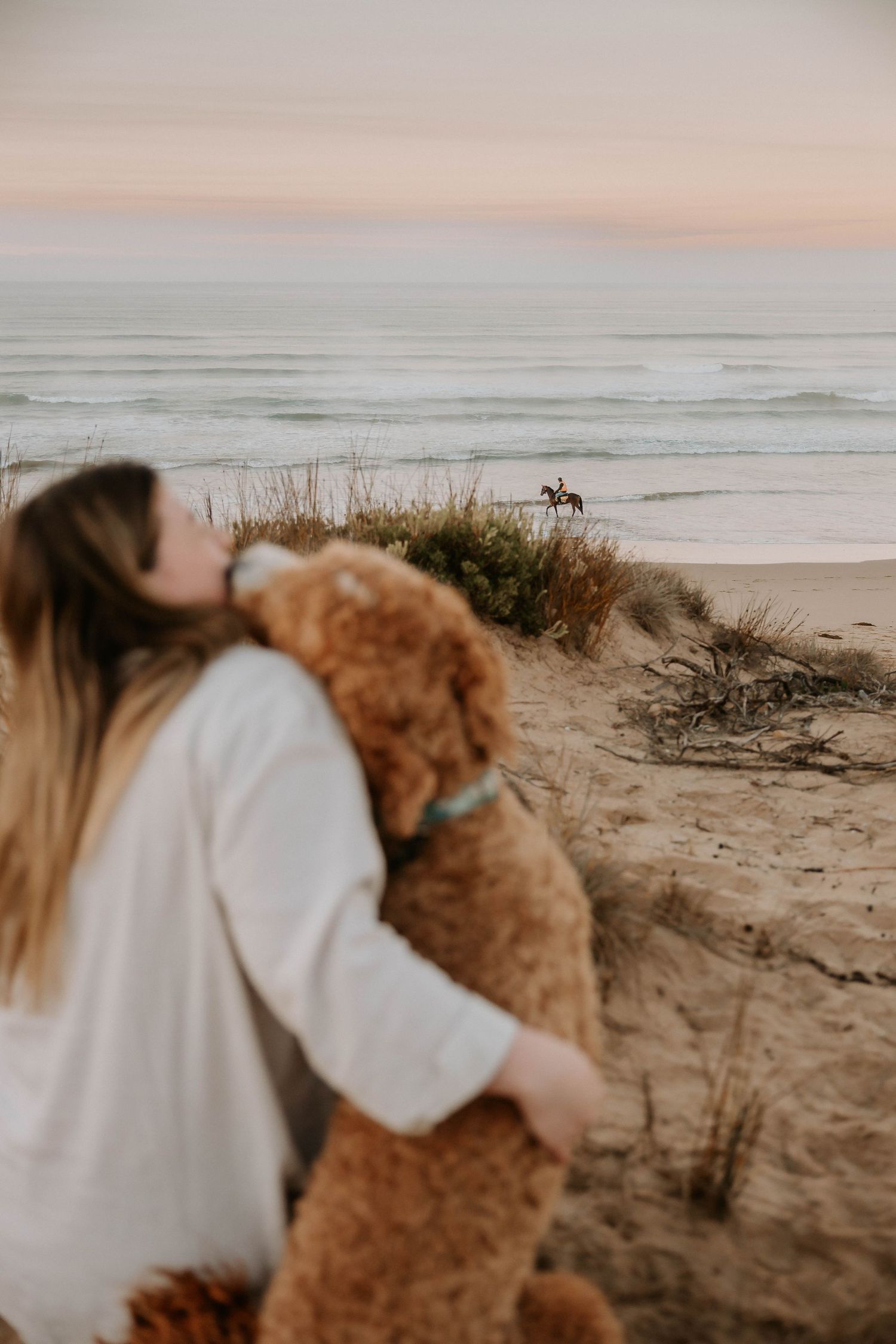 A person hugs a teddy bear while sitting on sandy dunes overlooking a beach with horse riders in the distance at sunset.