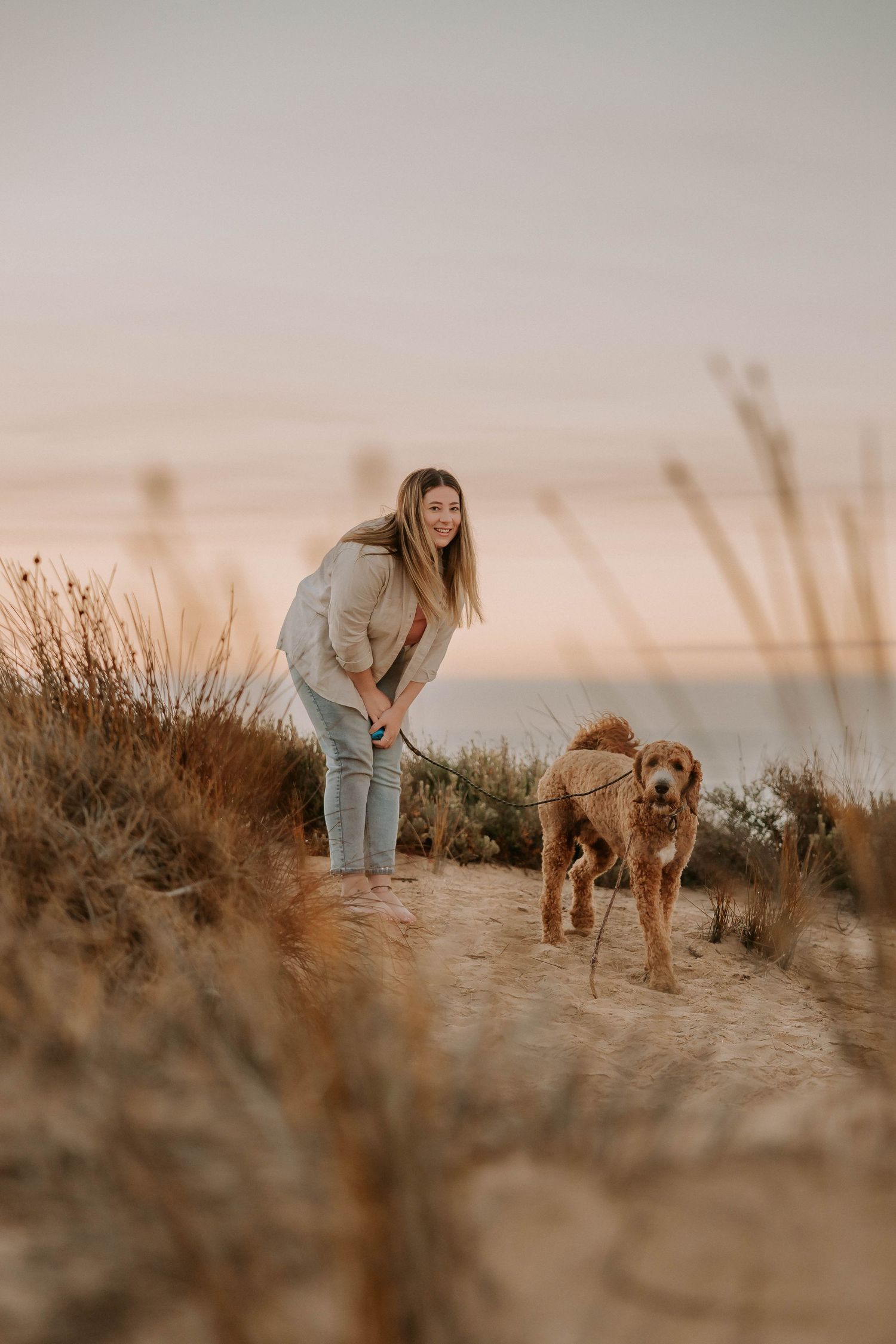 A person in casual clothing walks with their golden brown dog along a scenic coastal trail at sunset.