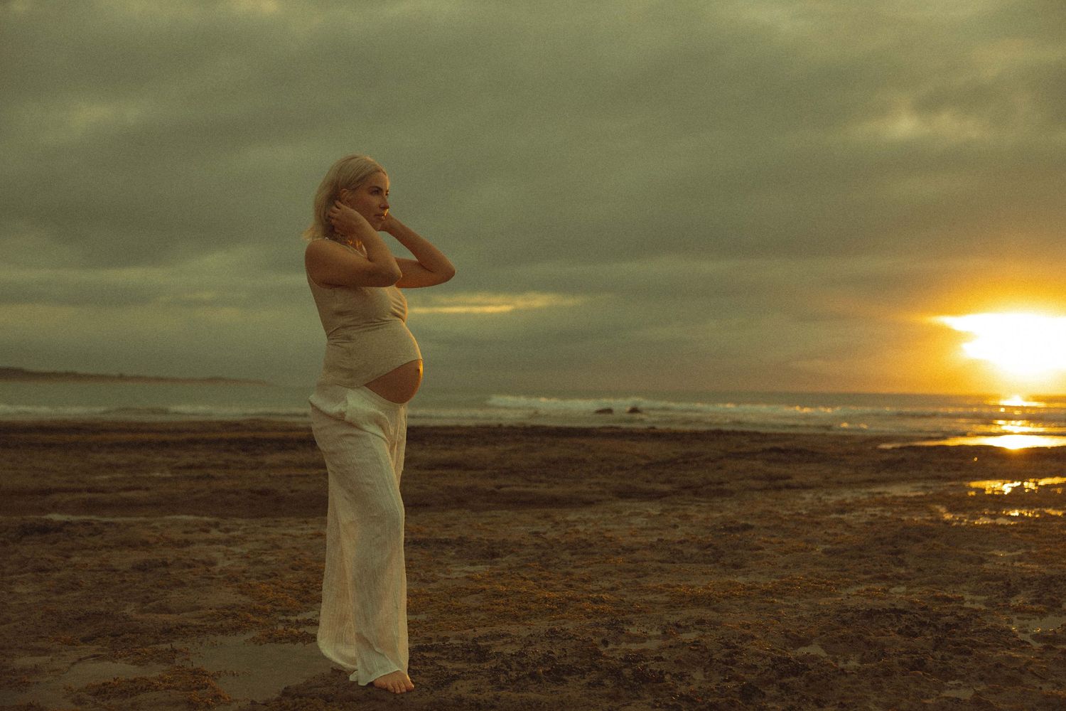 Figure in white stands on a beach at sunset with dramatic clouds and glowing sun reflecting on the ocean.