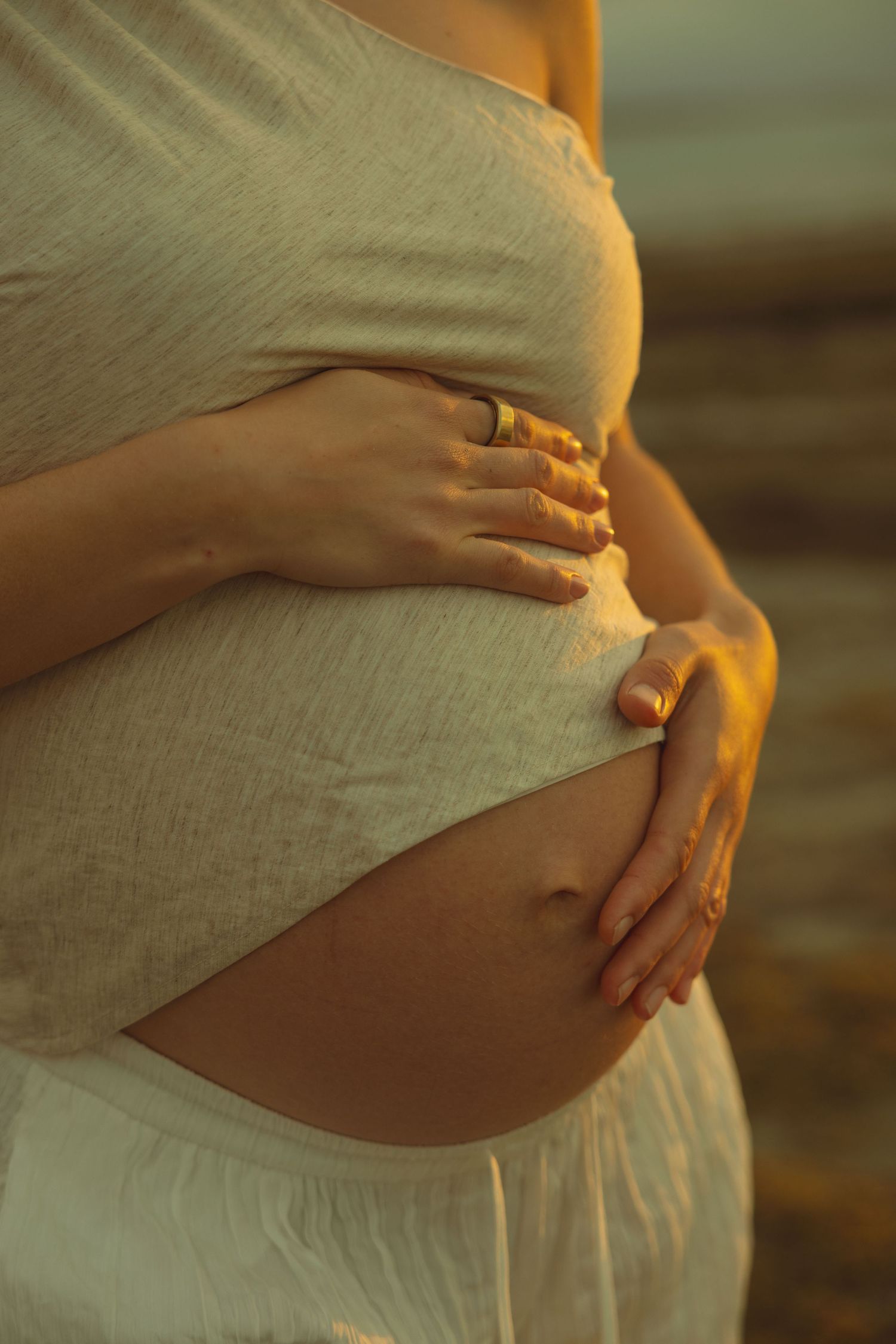 Close-up of pregnant belly in soft sunlight with hands tenderly cradling the bump, wearing white flowy fabric.