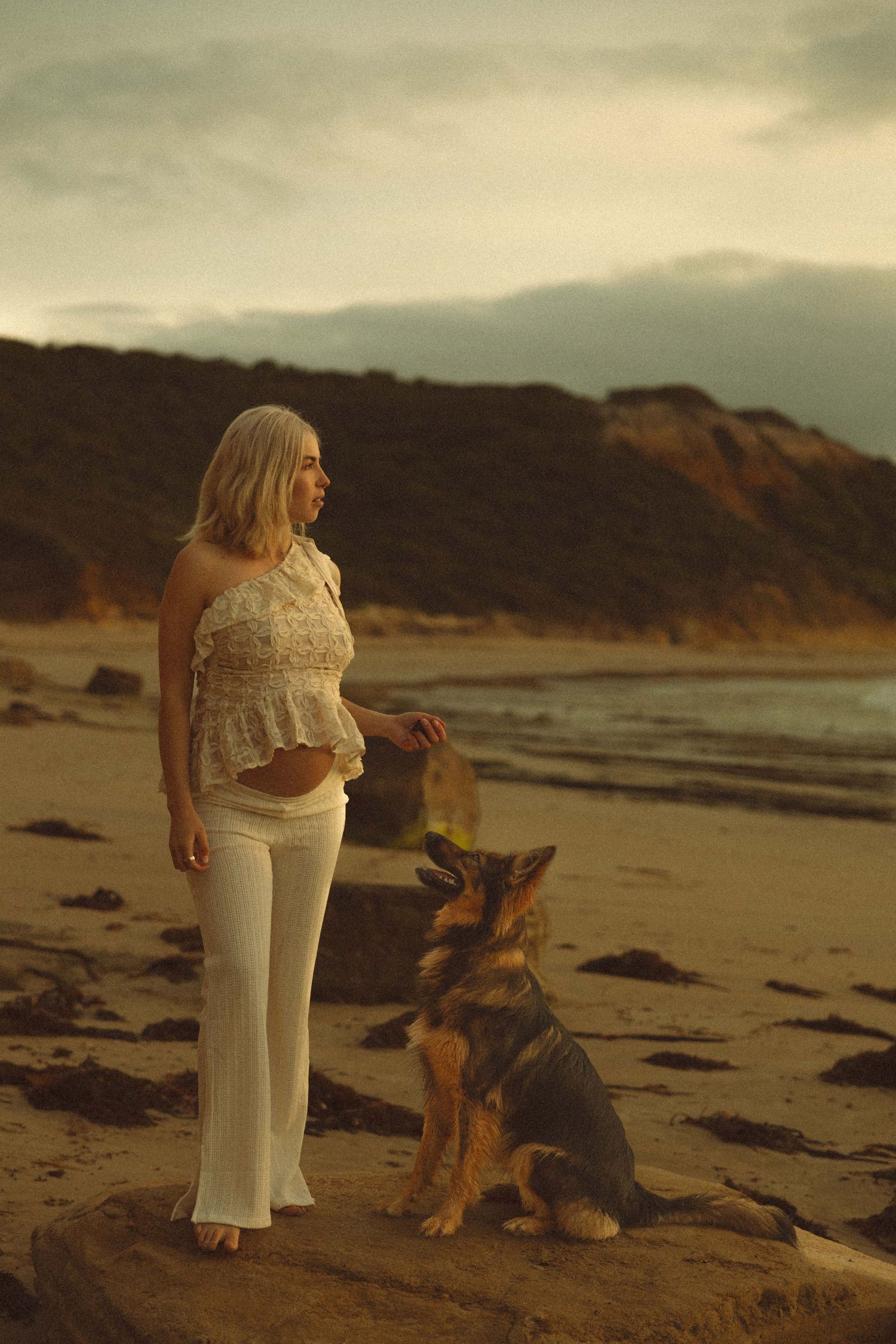 A German Shepherd sits attentively on a beach at sunset beside someone wearing white pants and a lace top.