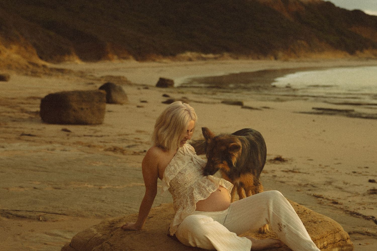 A person in white clothing sits on a sandy beach with their German Shepherd dog near coastal cliffs at sunset.