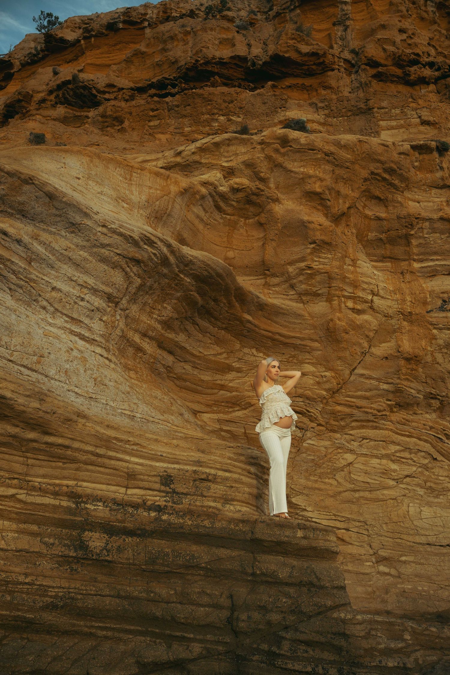 Someone in white stands on a dramatic layered sandstone cliff face against a moody sky.