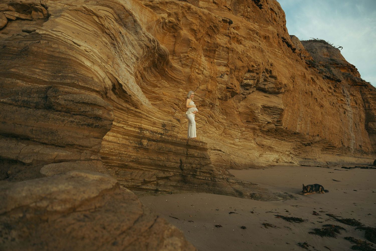 A lone figure stands against a dramatic sandstone cliff face on a sandy beach at dusk.