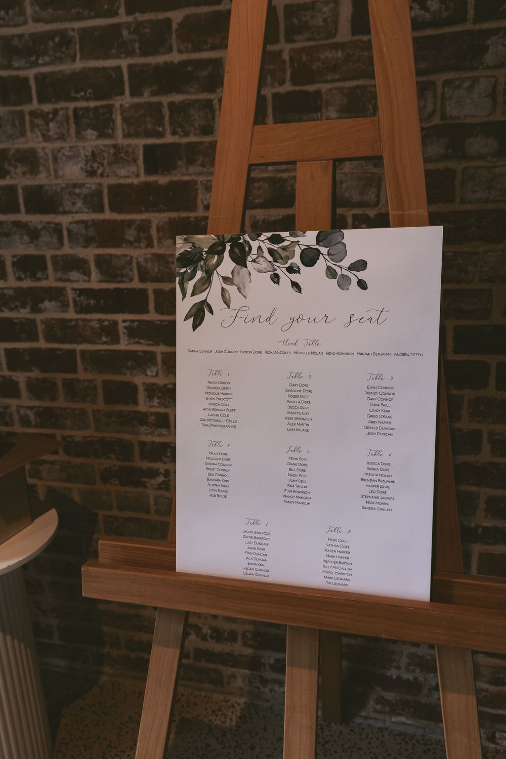 Wedding seating chart with floral design displayed on wooden easel against brick wall.