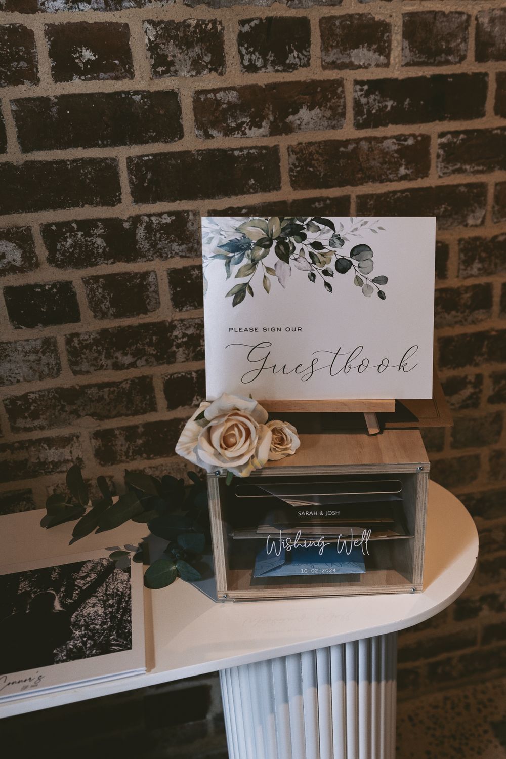 Wedding guestbook setup with rose decoration on white column near brick wall.