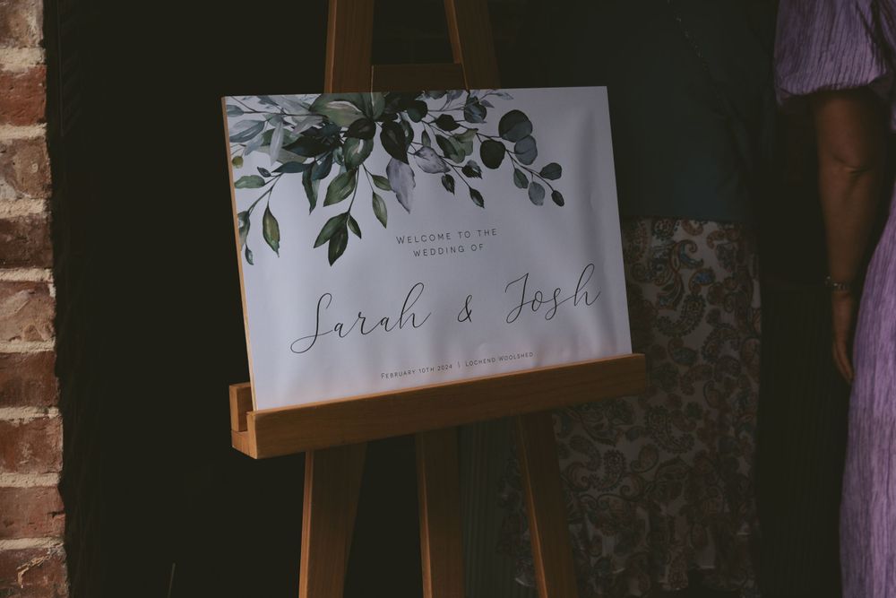 Elegant wedding signage with floral details displayed on a wooden easel against a rustic brick wall.