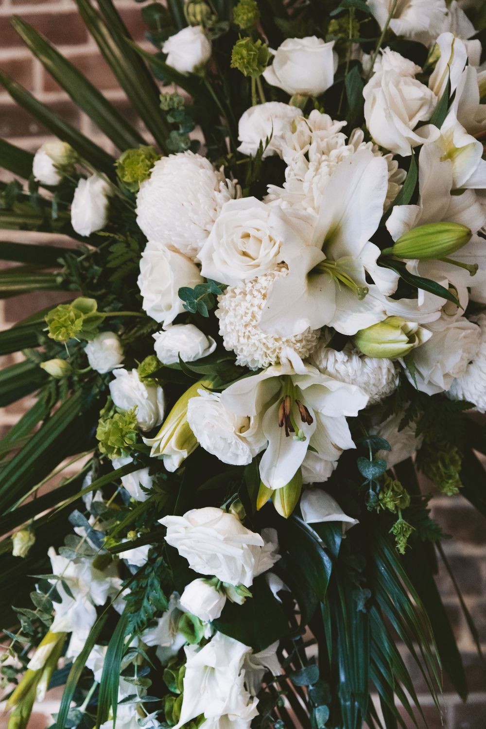 Luxurious white wedding bouquet featuring roses, lilies and greenery against a brick backdrop.