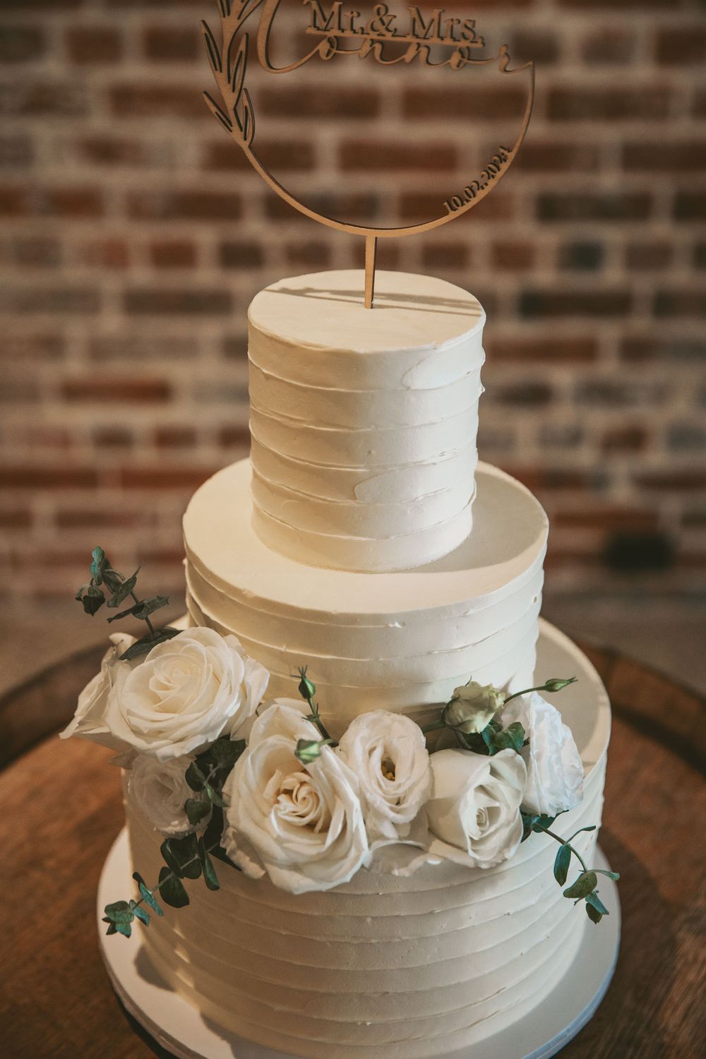 Three-tiered white wedding cake decorated with roses and a golden circular topper.